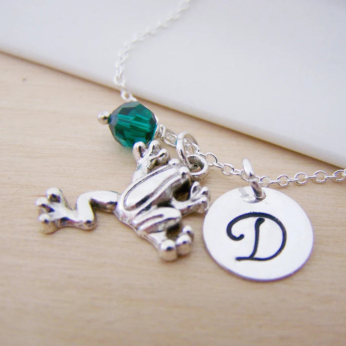 Necklace with silver frog charm, green bead, and round disc with letter 'D' on a light wooden surface.