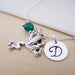 Necklace with silver frog charm, green bead, and round disc with letter 'D' on a light wooden surface.