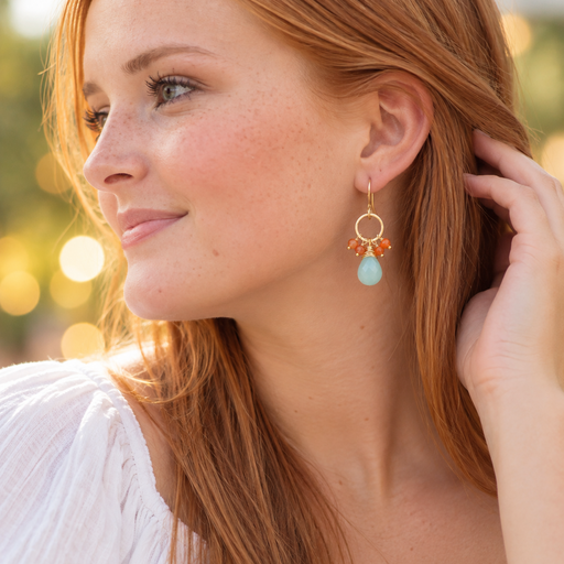 A close-up lifestyle shot of a woman with red hair wearing a gold dangle earring with a light blue teardrop stone and orange crystal accents.