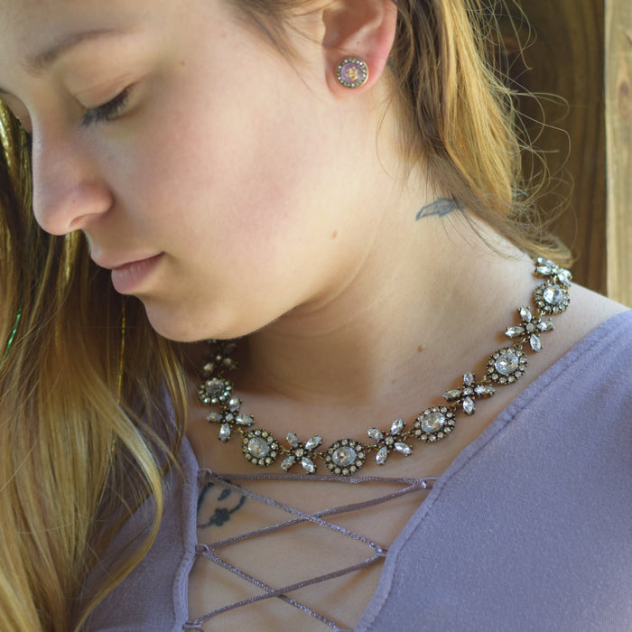 A close-up lifestyle shot of a woman wearing a silver-toned floral crystal necklace and matching stud earrings, wearing a lavender top.