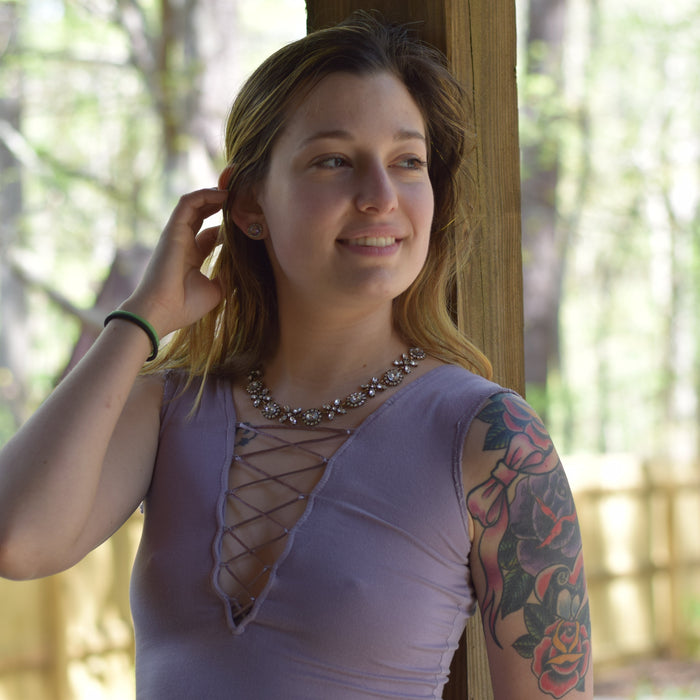 A woman with a floral tattoo on her left arm leans against a wooden post in an outdoor wooded setting. She is wearing a lavender sleeveless top with a lace-up front and a silver beaded necklace.