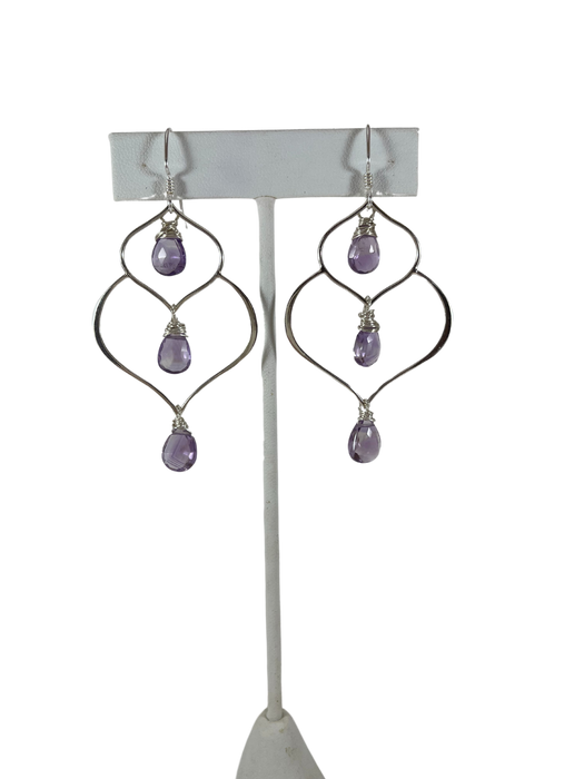 A pair of handcrafted silver and amethyst dangle earrings featuring a unique, organic wire frame design. Each earring has three faceted teardrop-shaped amethyst gemstones, wire-wrapped and suspended from delicate silver ear hooks. The background is a bright, clean white
