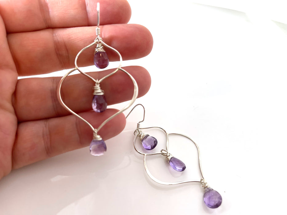 a hand holding a pair of earrings. A pair of handcrafted silver and amethyst dangle earrings featuring a unique, organic wire frame design. Each earring has three faceted teardrop-shaped amethyst gemstones, wire-wrapped and suspended from delicate silver ear hooks. The background is a bright, clean white