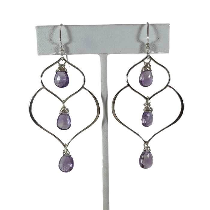 an earring stand holding a pair of earrings. A pair of handcrafted silver and amethyst dangle earrings featuring a unique, organic wire frame design. Each earring has three faceted teardrop-shaped amethyst gemstones, wire-wrapped and suspended from delicate silver ear hooks. 