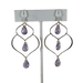 an earring stand holding a pair of earrings. A pair of handcrafted silver and amethyst dangle earrings featuring a unique, organic wire frame design. Each earring has three faceted teardrop-shaped amethyst gemstones, wire-wrapped and suspended from delicate silver ear hooks. 