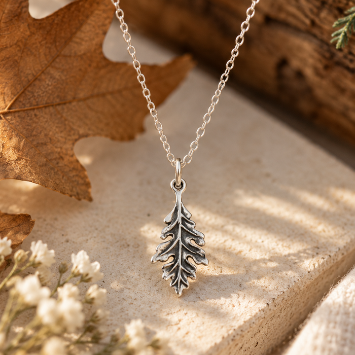 A silver leaf pendant necklace on a delicate silver chain, styled on a textured stone slab with dried autumn leaves and small white flowers.