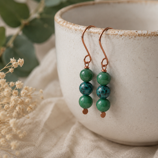 Green beaded earrings hanging in front of a white ceramic cup with green leaves and flowers in the background.