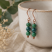 Green beaded earrings hanging in front of a white ceramic cup with green leaves and flowers in the background.