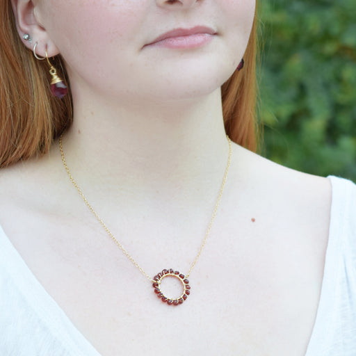 a gold necklace with a circle wire wrapped with garnet gemstones shown on a fair skinned model in white shirt with red hair
