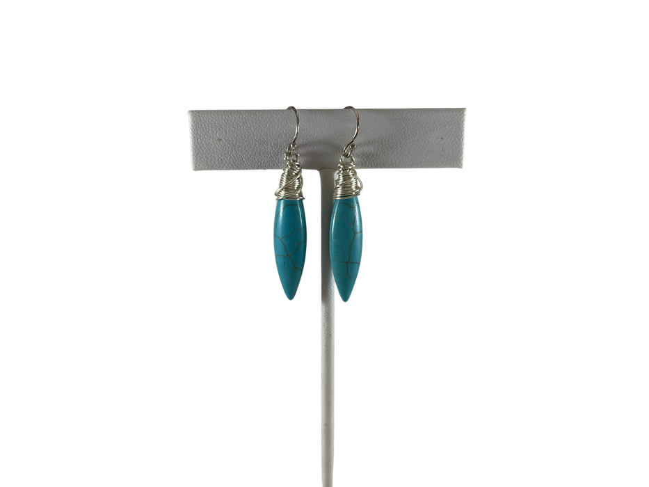 White leather earrings stand with earrings. The earrings feature elongated, marbled turquoise stones wrapped in delicate silver wire, suspended from silver ear hooks.