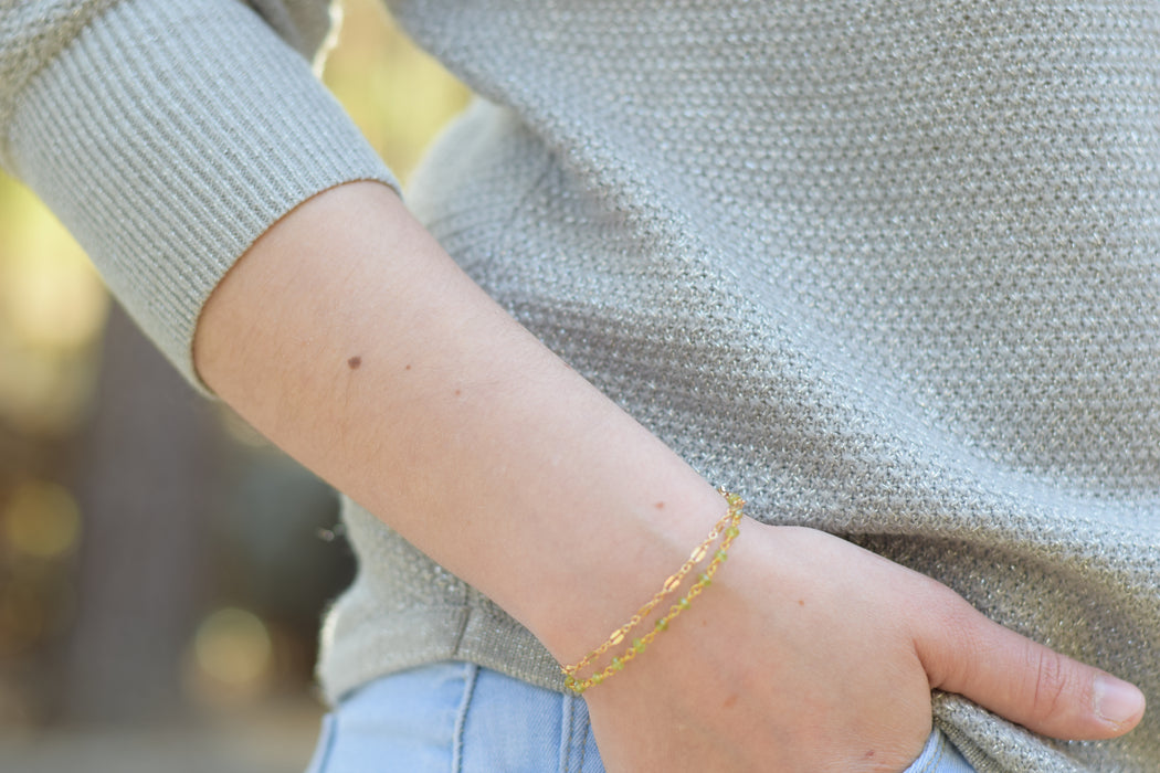 close up of a lady's wrist wear a dual chain bracelet with green peridot birthstones and gold chain.