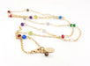 A delicate gold chain necklace adorned with small, faceted multicolored gemstone beads. The gemstones, including red, blue, green, purple, and clear, are evenly spaced along the chain, creating a vibrant and playful look. The necklace is displayed against a white background, with its gold clasp and a small engraved charm visible in the front, signed CY for CY Design Studio