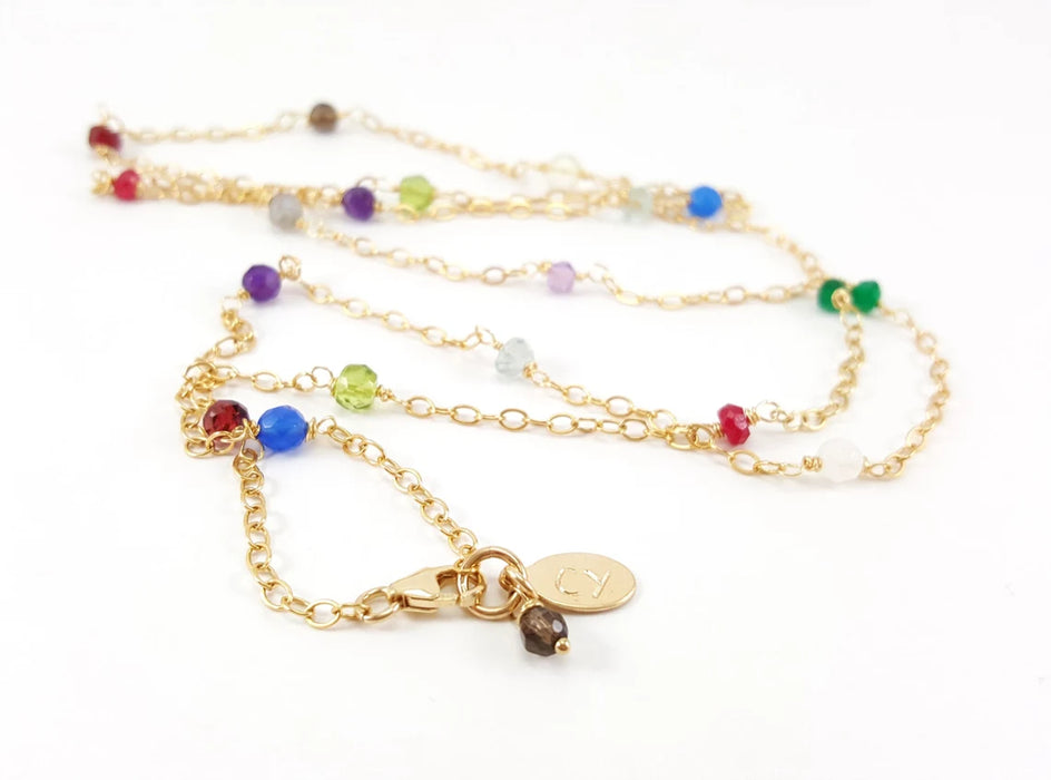 A delicate gold chain necklace adorned with small, faceted multicolored gemstone beads. The gemstones, including red, blue, green, purple, and clear, are evenly spaced along the chain, creating a vibrant and playful look. The necklace is displayed against a white background, with its gold clasp and a small engraved charm visible in the front, signed CY for CY Design Studio