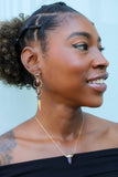 A close-up of a smiling woman with medium-dark skin tone wearing a black off-the-shoulder top. Her hair is styled in a natural curly updo with braided sections on the crown. She wears a statement earring with multiple gold hoops, a dangling gemstone, and a pointed charm, along with a delicate gold necklace featuring a pendant with a horn-shaped stone. She has a subtle winged eyeliner and a radiant complexion. The background is blurred with a light, neutral tone.