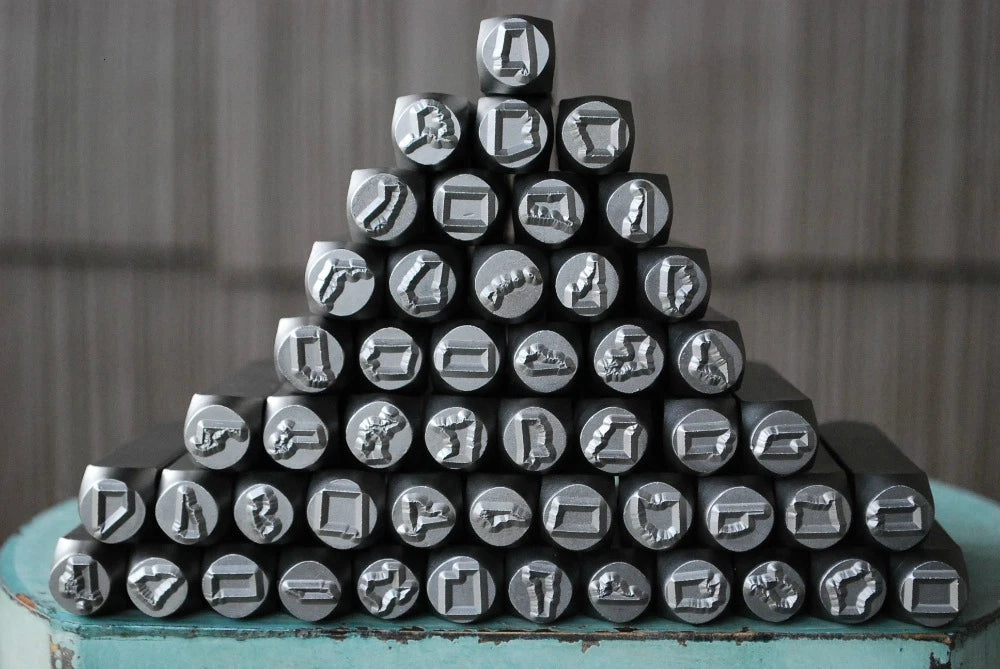 A pyramid arrangement of black metal letterpress printing stamps with various engraved symbols, displayed on a distressed teal wooden surface against a gray wooden background.
