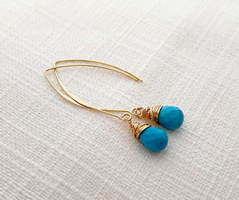 A pair of handmade gold and turquoise drop earrings featuring faceted turquoise gemstones wrapped in delicate gold wire. The earrings dangle from elegant gold-filled marquise-shaped ear wires. Displayed on a neutral textured fabric background.