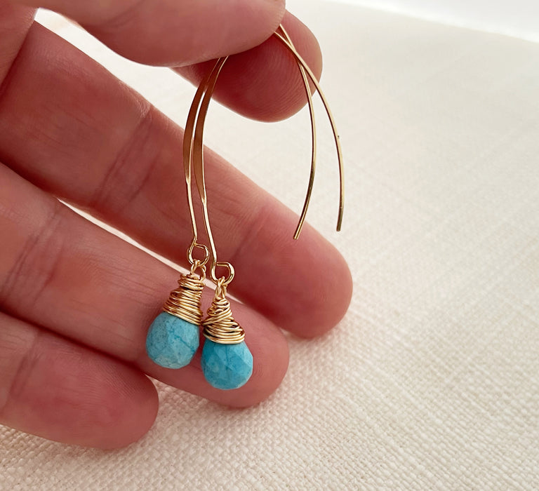 A hand holding a pair of earrings. A pair of handmade gold and turquoise drop earrings featuring faceted turquoise gemstones wrapped in delicate gold wire. The earrings dangle from elegant gold-filled marquise-shaped ear wires. Displayed on a neutral textured fabric background.