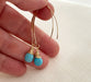 A hand holding a pair of earrings. A pair of handmade gold and turquoise drop earrings featuring faceted turquoise gemstones wrapped in delicate gold wire. The earrings dangle from elegant gold-filled marquise-shaped ear wires. Displayed on a neutral textured fabric background.