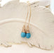 A pair of handmade gold and turquoise drop earrings featuring faceted turquoise gemstones wrapped in delicate gold wire. The earrings dangle from elegant gold-filled marquise-shaped ear wires. Displayed on a neutral feather textured background.