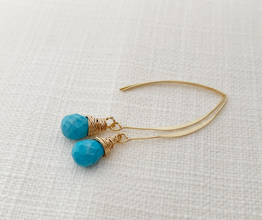 A pair of handmade gold and turquoise drop earrings featuring faceted turquoise gemstones wrapped in delicate gold wire. The earrings dangle from elegant gold-filled marquise-shaped ear wires. Displayed on a neutral textured fabric background.