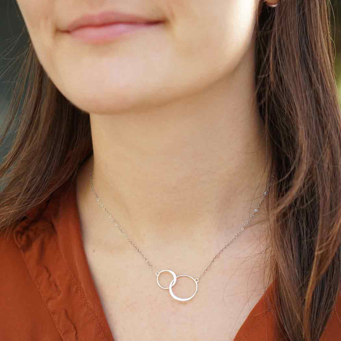 close-up of a woman wearing a sterling silver necklace with two interlocking circles. The delicate chain sits comfortably around her neck, and the minimalist pendant rests just above her collarbone. She is wearing a rust-colored blouse, and her long brown hair cascades over her shoulder.