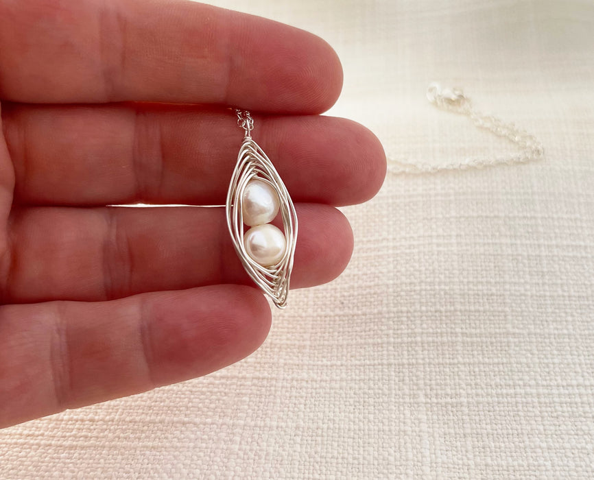 A handcrafted silver wire pendant featuring two white freshwater pearls nestled inside a delicate, swirling wire cage. The pendant is held between the fingers, showcasing its intricate design and petite size. A silver chain extends into the softly textured background, complementing the elegant and minimalist aesthetic