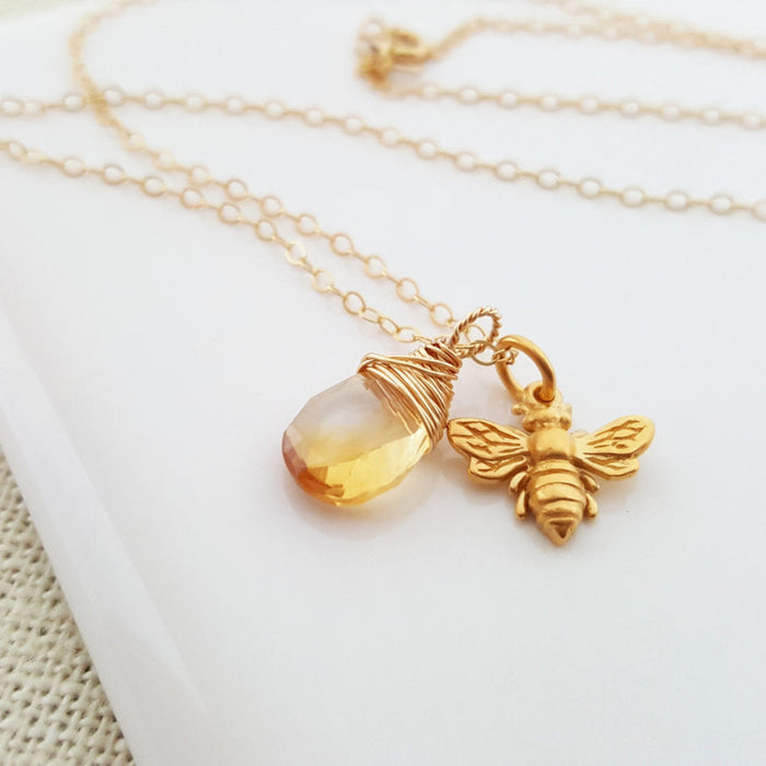 A 14k gold filled necklace with a detailed bee charm and a sparkling citrine briolette pendant, displayed on a white plate.